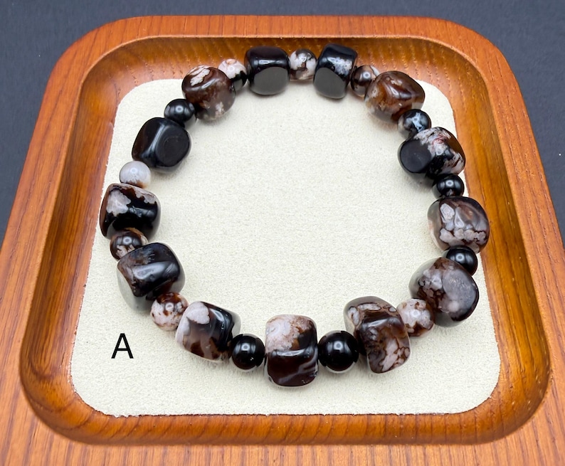 May include: A beaded bracelet featuring a mix of black, brown, and white stones. The bracelet is composed of various shapes and sizes of beads, including rounded and rectangular ones. The bracelet is displayed on a cream-colored surface inside a wooden tray.