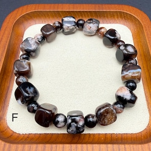 May include: A beaded bracelet featuring a variety of dark, polished stones in shades of brown, black, and gray. The bracelet is composed of both rounded and cube-shaped beads, strung together to form a circular design. The letter "F" is visible.