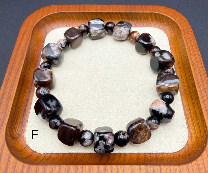 May include: A beaded bracelet featuring a variety of dark, polished stones in shades of brown, black, and gray. The bracelet is composed of both rounded and cube-shaped beads, strung together to form a circular design. The letter "F" is visible.