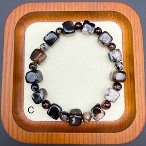 May include: A beaded bracelet featuring a mix of dark brown, black, and speckled gray stones. The bracelet includes both rounded and square-cut beads, creating a unique texture. The bracelet is displayed in a wooden tray.