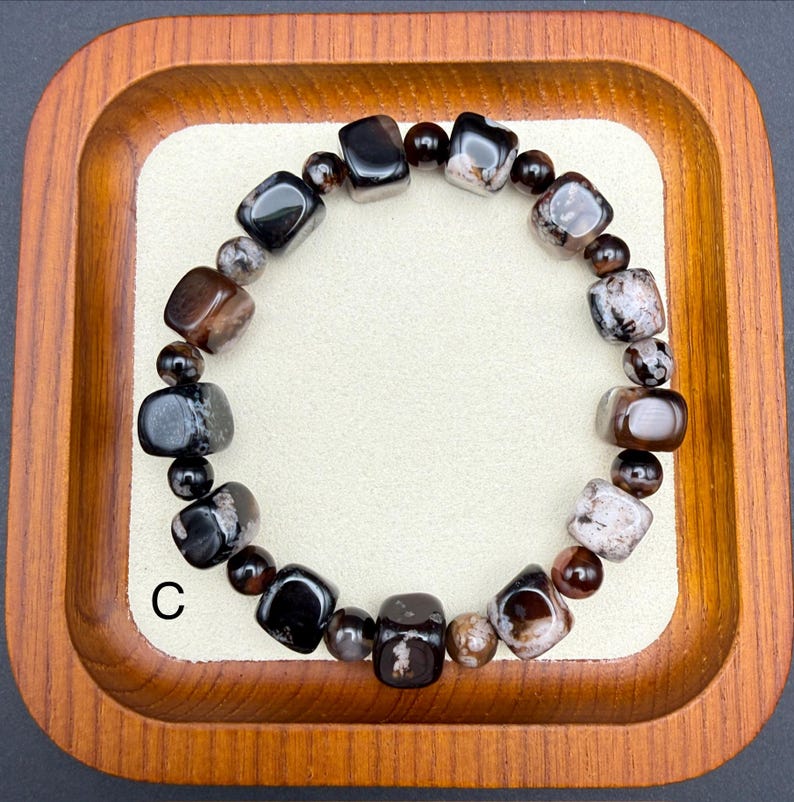 May include: A beaded bracelet featuring a mix of dark brown, black, and speckled gray stones. The bracelet includes both rounded and square-cut beads, creating a unique texture. The bracelet is displayed in a wooden tray.