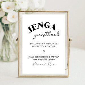Puede incluir: Un cartel blanco con texto negro que dice "JENGA guestbook" y "BUILDING NEW MEMORIES ONE BLOCK AT A TIME." Debajo del texto hay un pequeño símbolo de corazón y el texto "PLEASE SIGN A PIECE AND SHARE YOUR WELL WISHES FOR THE NEW Mr. and Mrs."