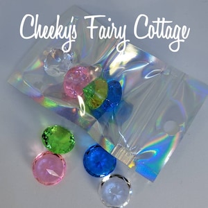 May include: A clear plastic bag with a zipper closure containing six iridescent gem-shaped decorations. The gems are in various colors, including clear, pink, green, blue, and yellow. The bag is on a white surface with the text "Cheeky's Fairy Cottage" written in black.