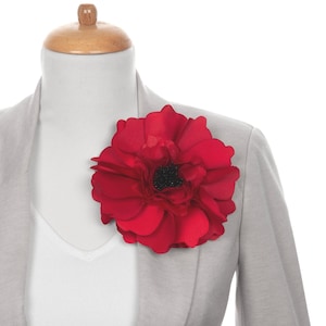 May include: A large, vibrant red flower brooch with a black center is pinned to a light gray blazer. The brooch is the focal point, adding a pop of color against the neutral jacket. The flower's petals are layered, creating depth.