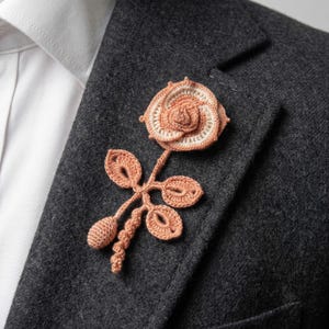 May include: A peach-colored crochet rose pin with a cream-colored center, attached to a stem with leaves and buds. The floral accessory is pinned to a dark gray blazer, paired with a white collared shirt.