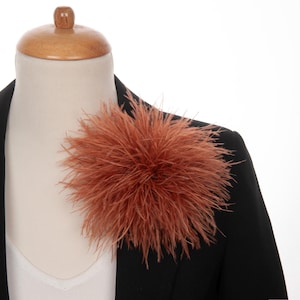 May include: A burnt orange feather brooch, affixed to a black blazer. The brooch is a large, round cluster of soft feathers. The blazer is worn over a white top, displayed on a mannequin.