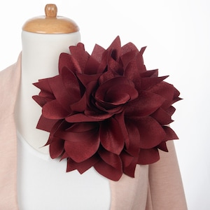 May include: A large, burgundy fabric flower brooch. The flower is made of multiple layers of petals and has a slightly ruffled texture.