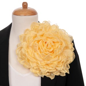 May include: A large, pale yellow fabric flower, resembling a peony, is pinned to a black blazer. The flower has multiple layers of delicate petals. The blazer is worn over a white top, displayed on a mannequin.