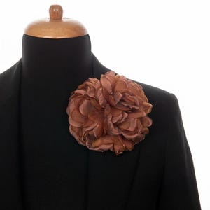 May include: A large, brown fabric flower pinned to the lapel of a black blazer. The flower has multiple layers of petals, creating a full, textured appearance. The blazer is displayed on a mannequin with a wooden head.