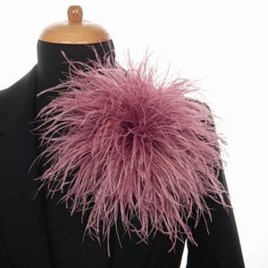 Może przedstawiać: Pierzasty dodatek w kolorze przydymionego różu, być może naramiennik lub broszka, przymocowany do czarnej marynarki. Pióra są długie i puszyste, tworząc obszerny, teksturowany efekt. Marynarka jest prezentowana na manekinie.
