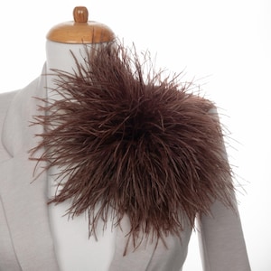 May include: A brown feather shoulder piece, possibly a decorative accessory, is shown on a mannequin wearing a light grey blazer. The feathers are dense and create a full, textured appearance. The background is plain white.