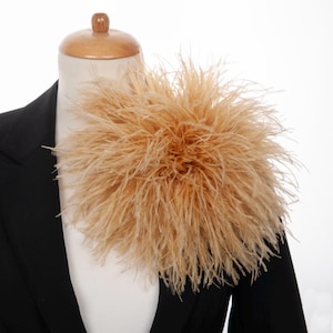 Può includere: Un grande accessorio rotondo in piume beige fissato su un blazer nero. L'accessorio è composto da numerose piume di struzzo sottili e leggere, disposte in un denso grappolo sferico che si irradia verso l'esterno partendo dal punto centrale.