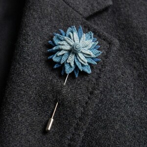 May include: A handmade, blue crochet flower lapel pin with a silver pin backing. The flower has multiple layers of blue and light blue petals, attached to a dark gray wool blazer.