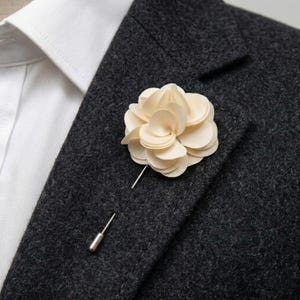 Könnte beinhalten: Ein cremefarbener Blumen-Revers-Anstecker mit silberner Nadel. Die Blume besteht aus geschichteten Blütenblättern und ist an einem dunkelgrauen Blazer über einem weißen Hemd befestigt.