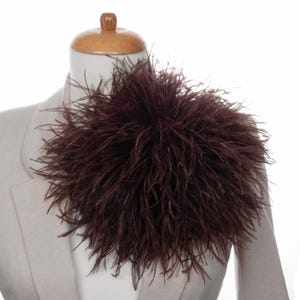 May include: A deep burgundy feather shoulder piece, possibly an accessory for formal wear, is shown on a mannequin. The feathers are densely packed, creating a full, textured look. The mannequin is wearing a light grey blazer.