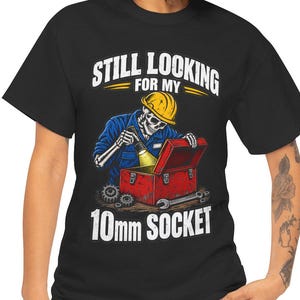 May include: A black t-shirt featuring a graphic of a skeleton wearing a yellow hard hat and blue work clothes, looking into a red toolbox with a flashlight. The text reads STILL LOOKING FOR MY 10mm SOCKET in white and yellow lettering.