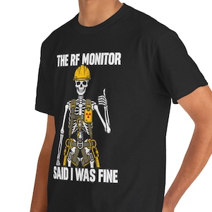 May include: A black short-sleeve t-shirt featuring a graphic of a skeleton wearing a yellow hard hat and a safety harness. The skeleton is giving a thumbs up. The text reads THE RF MONITOR SAID I WAS FINE in white capital letters.