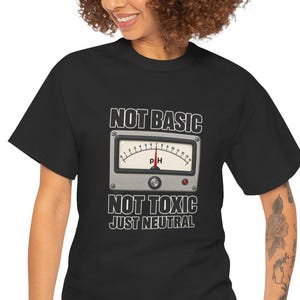 May include: A black short-sleeve t-shirt featuring a graphic of a pH meter gauge with a red needle pointing to the center. The text above the gauge reads NOT BASIC and the text below reads NOT TOXIC JUST NEUTRAL in white capital letters.