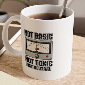 May include: A white ceramic mug featuring a graphic of a pH meter gauge with a red needle pointing to the center. The text above and below the gauge reads "NOT BASIC," "NOT TOXIC," and "JUST NEUTRAL" in bold, black, sans-serif capital letters.