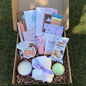 May include: A gift box filled with various items, including a candle, bath bomb, Tazo tea, Milano cookies, stickers, a notebook, and a small white towel tied with a purple ribbon. The box is lined with brown crinkle paper and sits on grass.