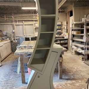 May include: A white curved bookshelf with five shelves and a cabinet door. The bookshelf is in a workshop setting.