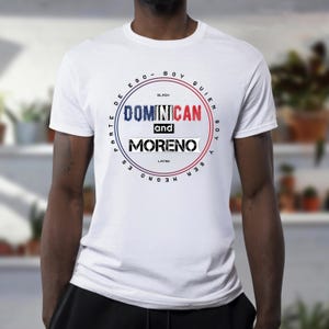 May include: White t-shirt with a circular graphic design. The design features the words "Dominican and Moreno" in a bold font, with additional text in a circular pattern around the central text. The design incorporates red, white, and blue colors.