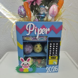 May include: A blue Easter-themed decorative box with the name "Piper" on the front. It features a bunny, eggs, and a chick. The top is decorated with pink flowers, carrots, and Easter eggs. The year "2026" is displayed on the front.
