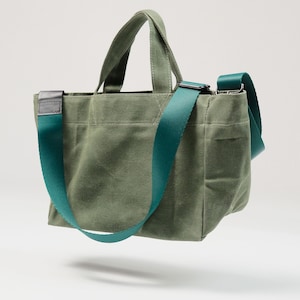 Può includere: Borsa tote verde oliva con una tracolla regolabile e manici abbinati verde acqua. La borsa ha una forma rettangolare con una piccola tasca sul davanti. La borsa è realizzata in resistente tela.