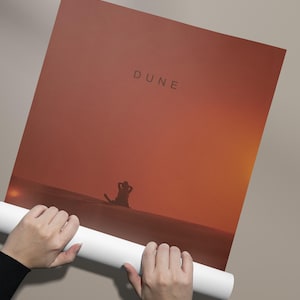 May include: A rolled up poster with a sunset background and the word "DUNE" in black text. A silhouette of a person is sitting in the sand in the foreground.