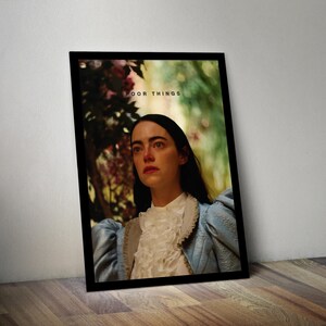 May include: A framed poster with a woman in a blue and white dress looking off to the side. The poster has the text "Poor Things" in black letters.