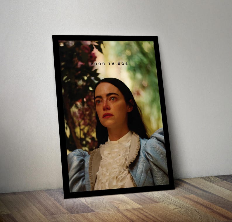 May include: A framed poster with a woman in a blue and white dress looking off to the side. The poster has the text "Poor Things" in black letters.