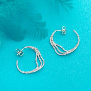 Peut inclure: Deux boucles d'oreilles argentées en forme de créoles, avec un design ajouré et délicat. Chaque boucle est ornée de petites pierres scintillantes. Elles sont présentées sur un fond turquoise vif, avec une plume douce en haut à gauche.