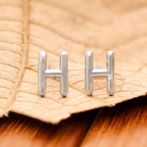Puede incluir: Un par de pendientes de botón plateados con la letra "H". Los pendientes se muestran sobre una hoja marrón contra un fondo de madera borroso. Los pendientes son pequeños y tienen un diseño simple y minimalista.