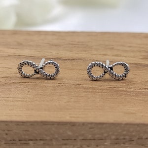 Pode incluir: Um par de brincos de prata em forma de símbolo de infinito. Os brincos são feitos de um design trançado.