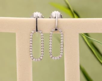 May include: A pair of silver-toned oval drop earrings. Each earring is outlined with small, clear gemstones. The earrings are displayed on a neutral-colored stand against a soft green background. The earrings have post backs.