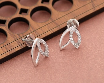 May include: A pair of silver stud earrings with a leaf design. Each earring features a curved, open leaf shape with a central oval filled with small, clear gemstones. The earrings are displayed on a pink surface with a wooden background.