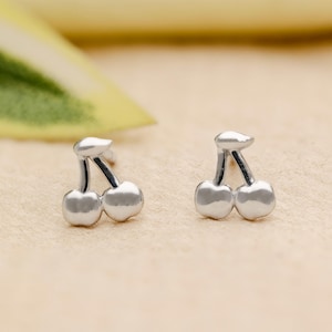 May include: A pair of silver-colored cherry stud earrings. Each earring features two round cherry shapes connected by a stem and a leaf-like detail. The earrings are displayed on a light beige surface.