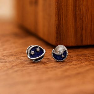 May include: A pair of silver-toned stud earrings featuring celestial motifs. One earring is shaped like a planet with a ring and two small stars, while the other is a crescent moon with a single star. Both have a deep blue, textured enamel inlay.