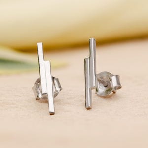 May include: A pair of silver stud earrings. Each earring has a vertical bar design with a stepped, geometric shape. The earrings are presented on a neutral, textured surface.