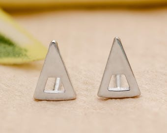 May include: A pair of silver triangle stud earrings. Each earring features a geometric triangle shape with a rectangular cutout in the center. The earrings are displayed on a light brown surface.