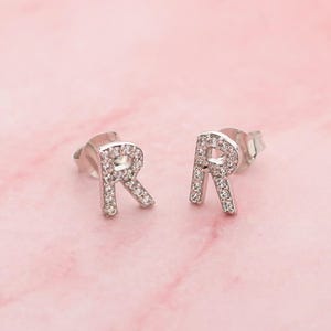 May include: A pair of silver stud earrings in the shape of the letter "R". Each earring is set with small, clear gemstones. The earrings are presented against a pink backdrop.