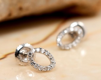 May include: A pair of silver oval stud earrings with small, clear gemstones set around the oval. The earrings are displayed on a light-colored, reflective surface.