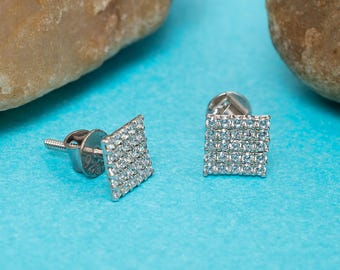 May include: Square-shaped stud earrings with a silver backing and a surface covered in small, clear gemstones. The earrings are displayed on a light blue surface, with two stones in the background.