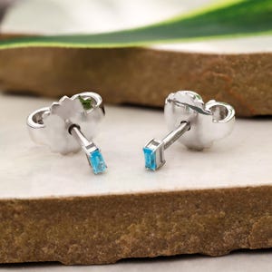 Puede incluir: Dos pendientes de plata con piedras rectangulares de color azul claro. Los pendientes se muestran sobre una superficie clara con un fondo texturizado. Los pendientes tienen un cierre de poste y mariposa.