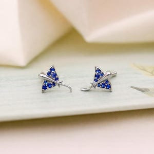 May include: A pair of silver-toned stud earrings shaped like stingrays, each featuring a metallic body encrusted with small, vibrant blue circular stones that cover the wings and central portion of the aquatic animal design.