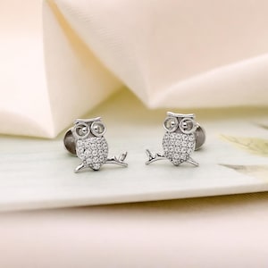 May include: A pair of silver-toned stud earrings shaped like owls perched on branches. Each owl features two circular eyes and a body encrusted with numerous small, clear, sparkling stones that catch the light, creating a bright, reflective surface.