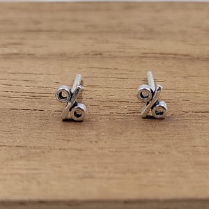 Puede incluir: Un par de pequeños pendientes de botón plateados con forma de símbolo de porcentaje. Los pendientes están colocados sobre una superficie de madera marrón claro.