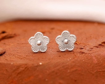 May include: A pair of silver-colored flower stud earrings. Each earring features a five-petal design with a textured surface and a small, round center. The earrings are displayed on a reddish-brown surface.