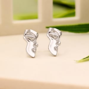 May include: A pair of silver earrings featuring a stylized face design. The earrings are small and delicate, perfect for everyday wear.