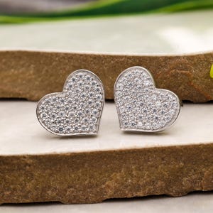 May include: A pair of heart-shaped stud earrings. Each earring features a silver-colored frame filled with small, sparkling, clear stones. The earrings are displayed on a light-colored surface with a brown, textured background.